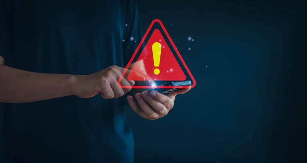 Manos de una persona interactuando con un teléfono móvil en un entorno oscuro, del cual emerge un gran ícono holográfico de triángulo de advertencia rojo y amarillo brillante, ilustrando la recepción de alertas de seguridad en campamentos mineros.